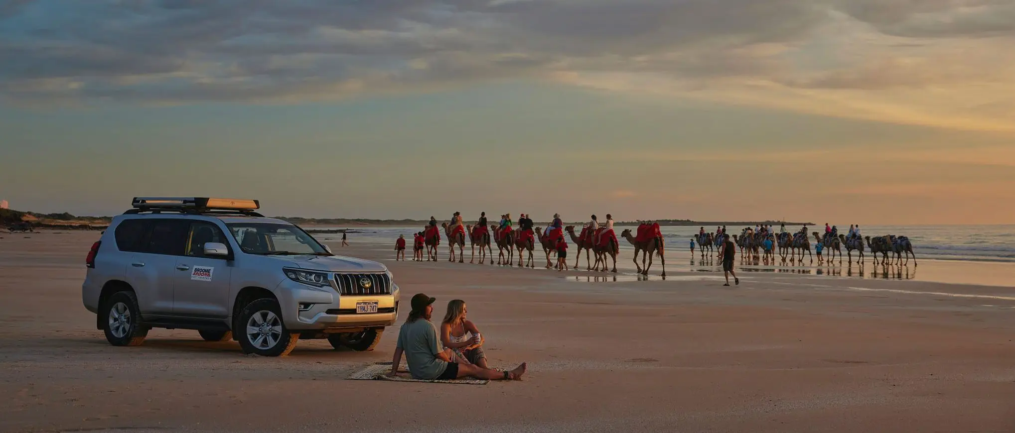 Couple watching the sunset on a beach with people riding camels in the background