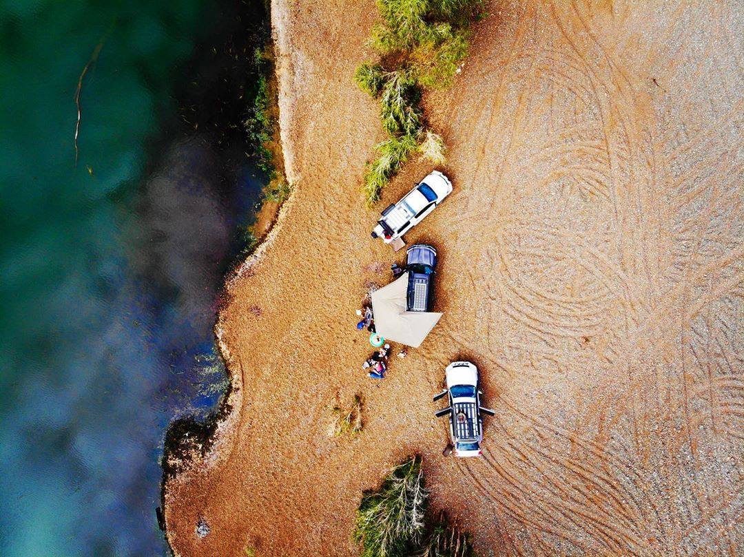 Three off road vehicles parked by a river with a camping setup