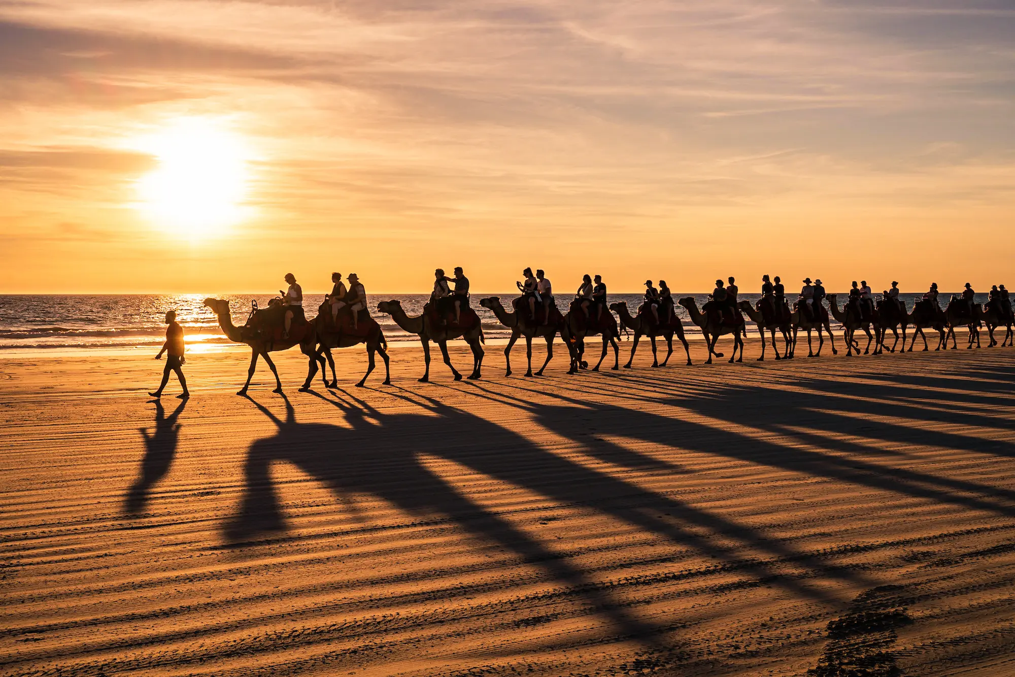People riding camels in formation on a beach as the sun sets